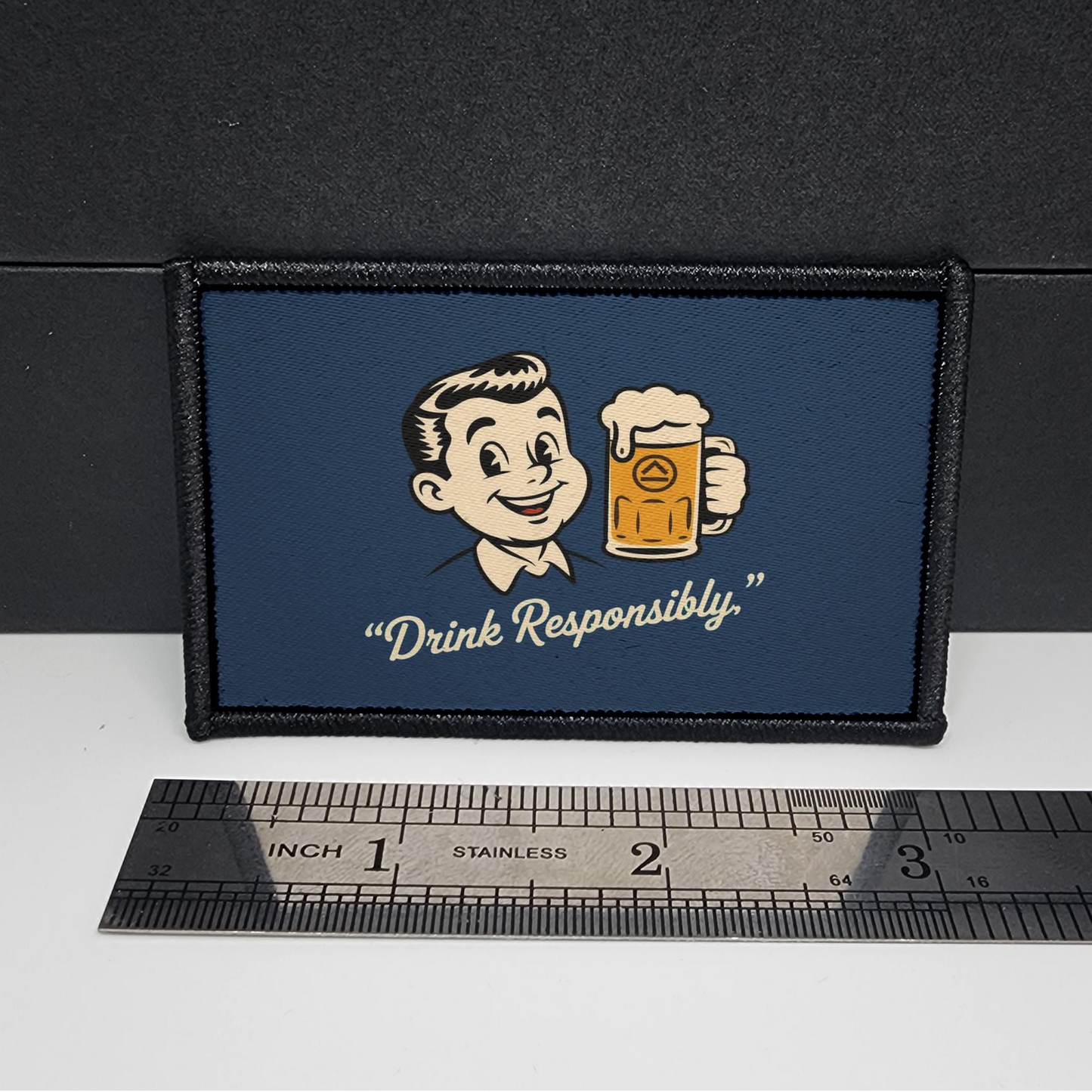 Patch with cartoon character holding a beer mug and 'Drink Responsibly' text on a blue background, placed on a white surface with a ruler for scale.
