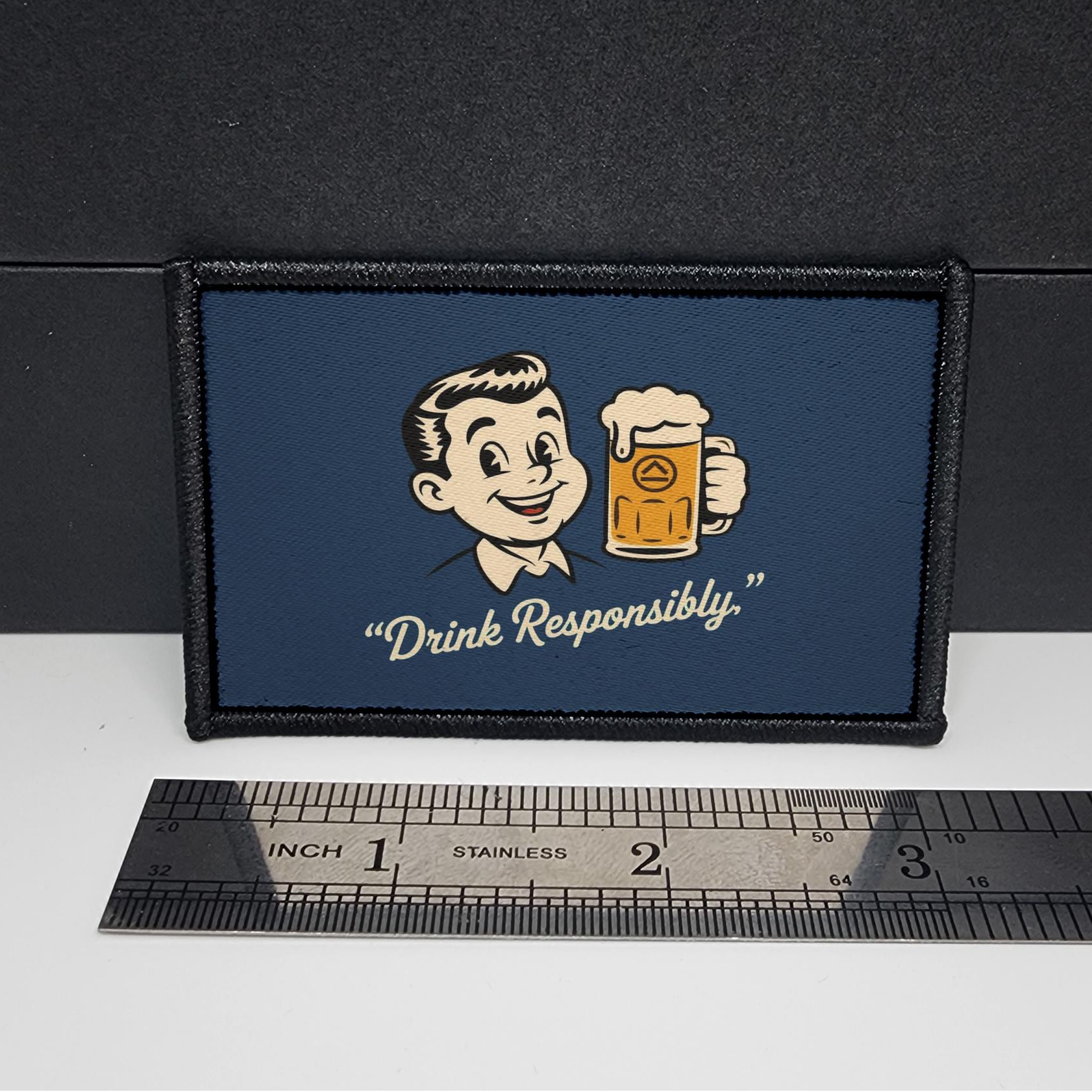 Patch with cartoon character holding a beer mug and 'Drink Responsibly' text on a blue background, placed on a white surface with a ruler for scale.