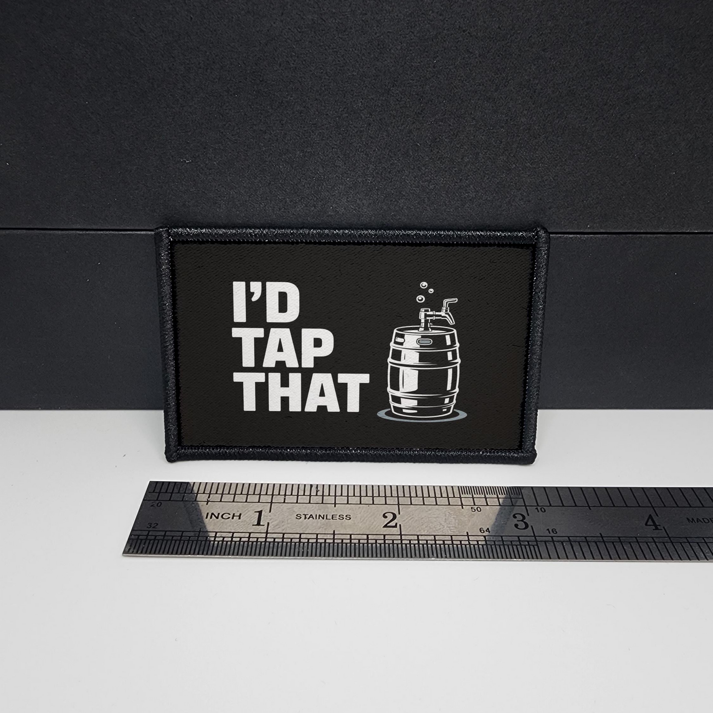 Black patch with 'I'd Tap That' text and a barrel graphic on a white surface with a ruler for scale.