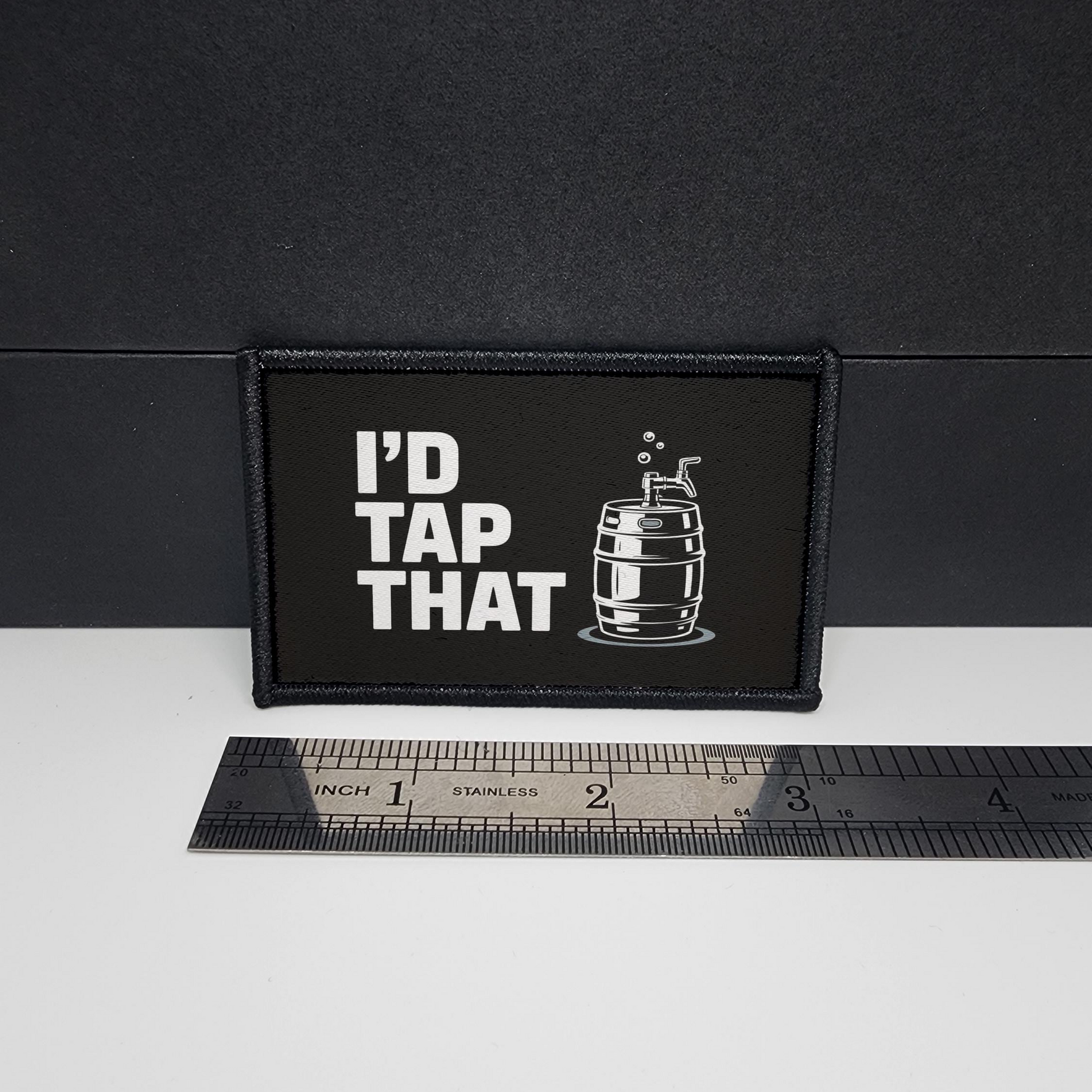 Black patch with 'I'd Tap That' text and a barrel graphic on a white surface with a ruler for scale.