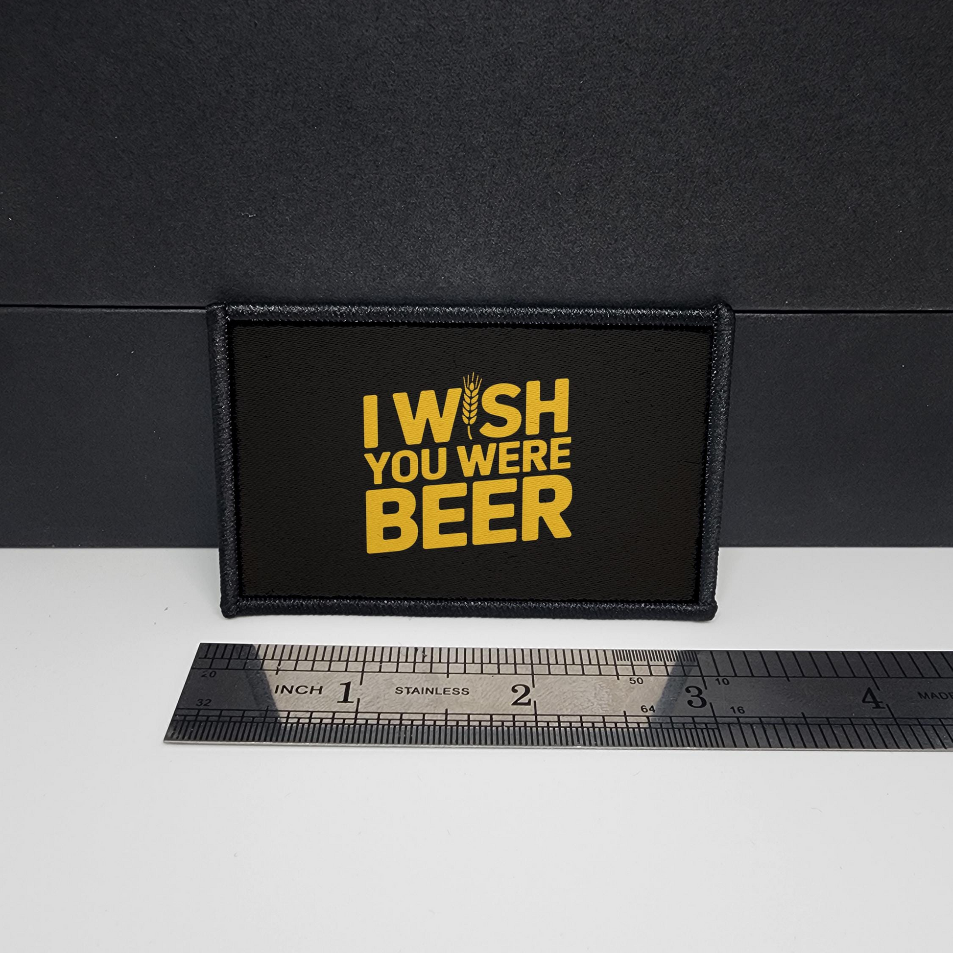 Black patch with yellow text 'I Wish You Were Beer' on a white surface with a ruler for scale.