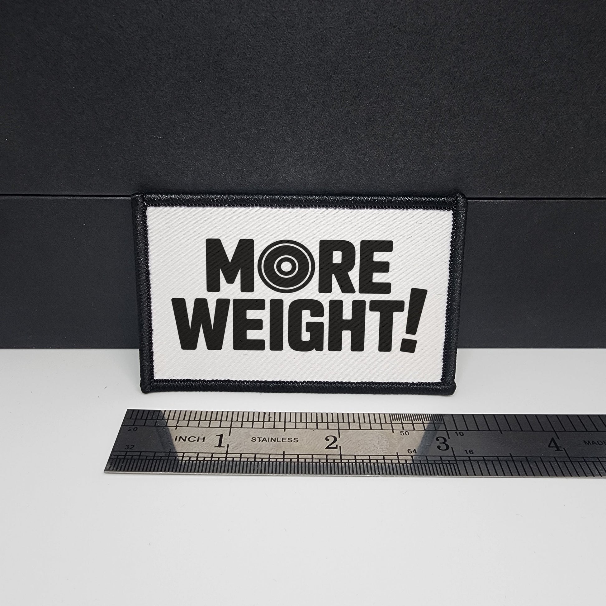 Patch with 'MORE WEIGHT!' text on a black background with a ruler for scale.