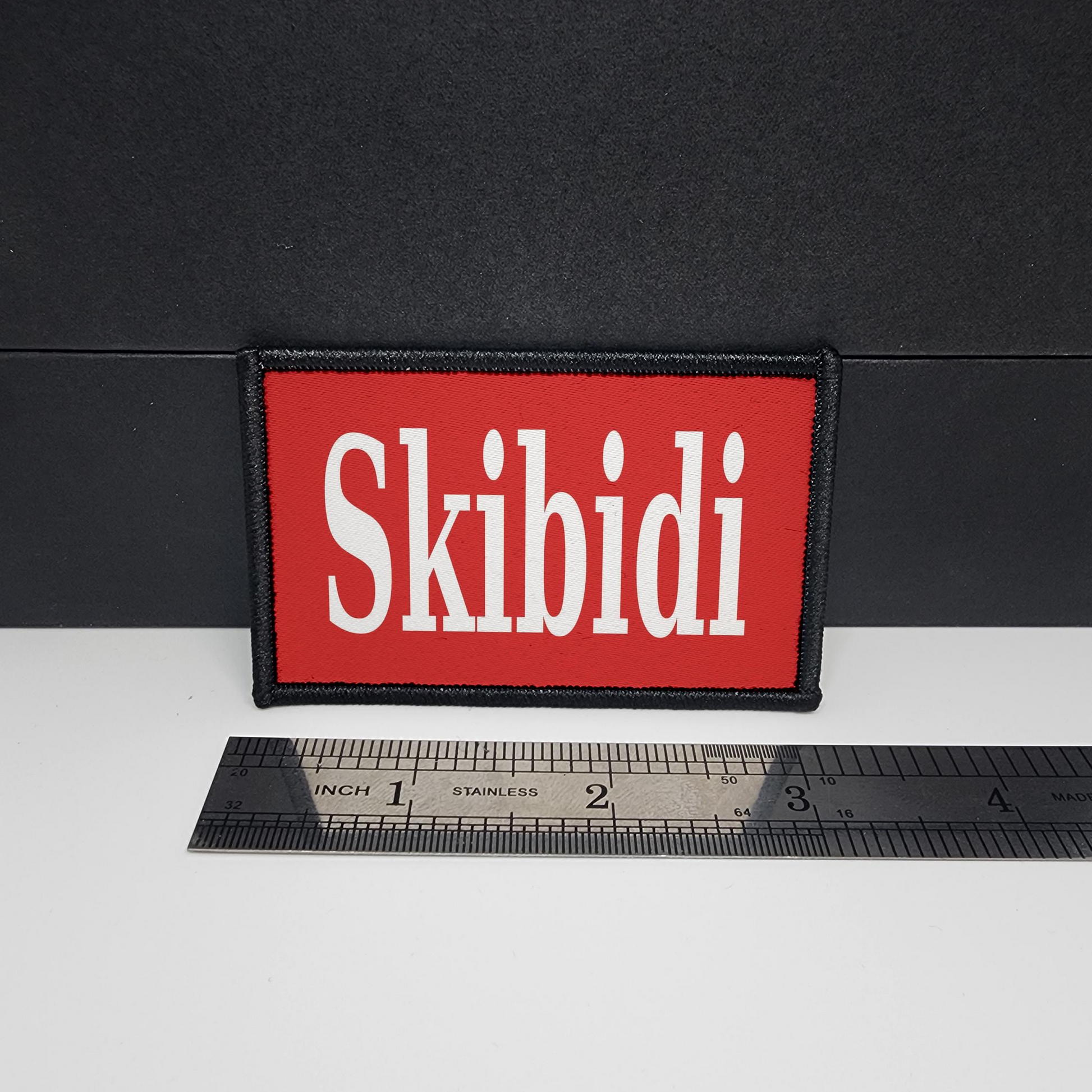 Red 'Skibidi' patch on a black background with a ruler for scale.

CAPS LOCK ORIGINAL - morale patch - 2x3 inch – hook and loop - iron-on - interchangeable patch - hat - bag